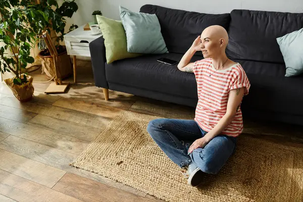 A beautiful bald woman reflects while sitting cross legged on a woven rug at home. — Stock Photo