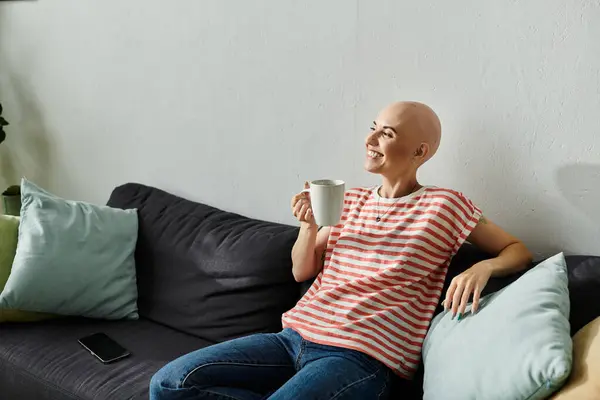 A bald woman smiles while sipping tea, relaxed on a comfortable couch amidst cozy decor. — Stock Photo