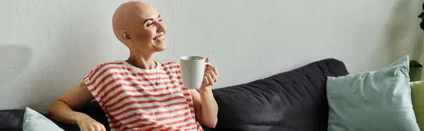 A happy bald woman enjoys her coffee while relaxing on a comfortable sofa filled with cushions. — Stock Photo