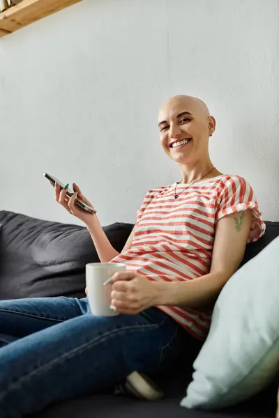 Eine lächelnde Frau mit Glatze entspannt sich auf der Couch, hält eine Tasse in der Hand und blättert auf ihrem Handy.. — Stockfoto