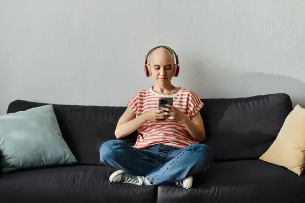A young, bald woman sits comfortably on a couch, lost in her music with headphones on. — Stock Photo