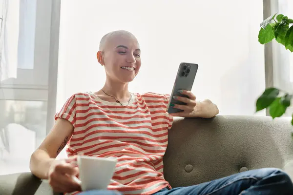 Una donna calva con alopecia sorride dolcemente mentre si rilassa con una tazza di caffè e il suo telefono. — Foto stock