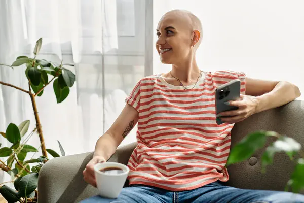 The woman with alopecia smiles while holding her phone and a cup of coffee in a cozy space. — Stock Photo