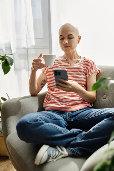 Una joven con alopecia bebe café y comprueba su teléfono en un espacio acogedor y luminoso. - foto de stock