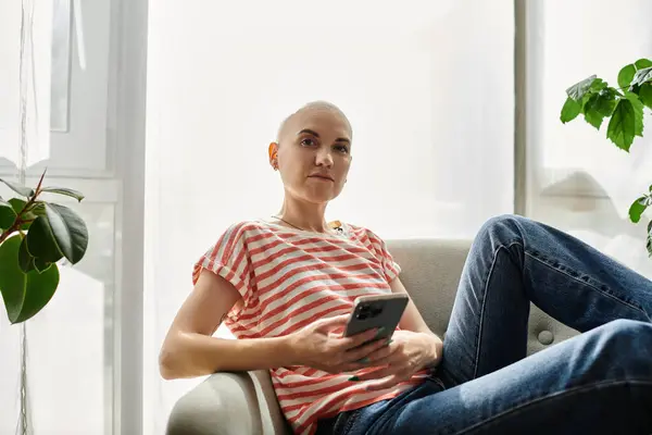 Eine junge Frau mit Glatze und Haarausfall sitzt gemütlich auf einem Sofa und greift mit ihrem Smartphone ein. — Stockfoto