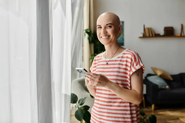 Eine junge Frau mit Alopezie steht am Fenster und lächelt strahlend, während sie mit ihrem Handy interagiert. — Stockfoto