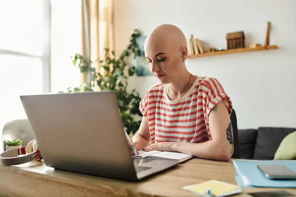 A young bald woman thoughtfully writes in her notebook while focused on her laptop. — Stock Photo