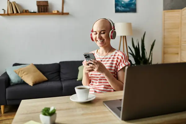 A joyful bald woman engages with her phone at a cozy home workspace, sipping coffee. — Stock Photo