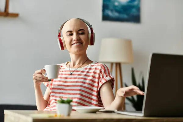 Una allegra donna calva siede alla scrivania, sorseggiando caffè e ascoltando musica sulle cuffie. — Foto stock