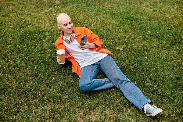 Una joven calva sonríe, sorbiendo café y usando su teléfono mientras está acostada sobre hierba verde. - foto de stock