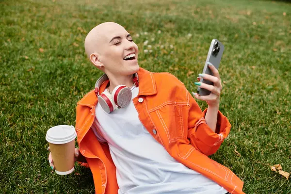 A joyful bald woman relaxes on the grass, sipping coffee and smiling at her phone. — Stock Photo