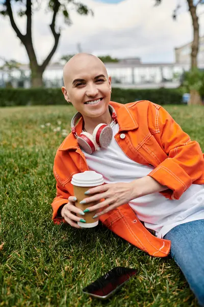 A joyful bald woman sits on the grass, sipping coffee and soaking up the sun. — Stock Photo
