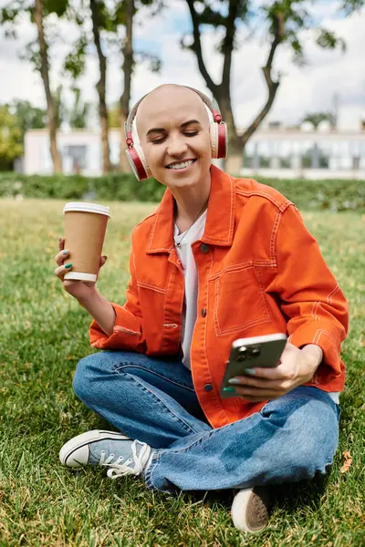 Una donna calva allegra si rilassa sull'erba con un caffè e cuffie, godendo la sua giornata. — Foto stock