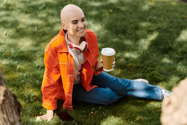 Eine fröhliche junge Frau mit Alopezie sitzt im Gras und nippt am Kaffee und sonnt sich im Sonnenlicht. — Stockfoto