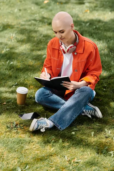 Une belle jeune femme avec alopécie s'assoit sur l'herbe, écrivant joyeusement dans son journal. — Photo de stock