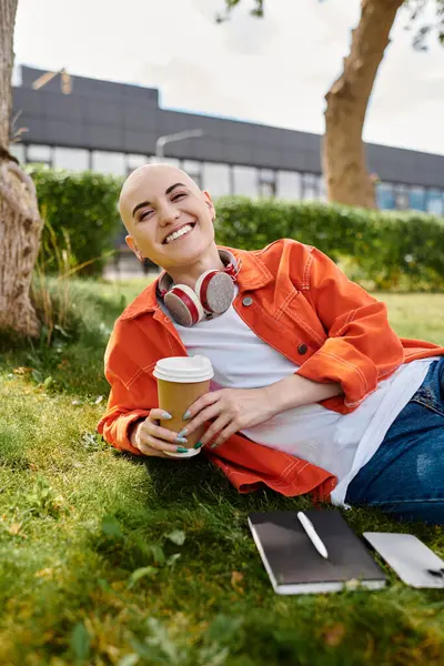 A young bald woman relaxes on the grass, smiling brightly with a cup of coffee in hand. — Stock Photo