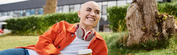 A young woman with a bald head smiles joyfully while relaxing on the grass, sipping coffee. — Stock Photo