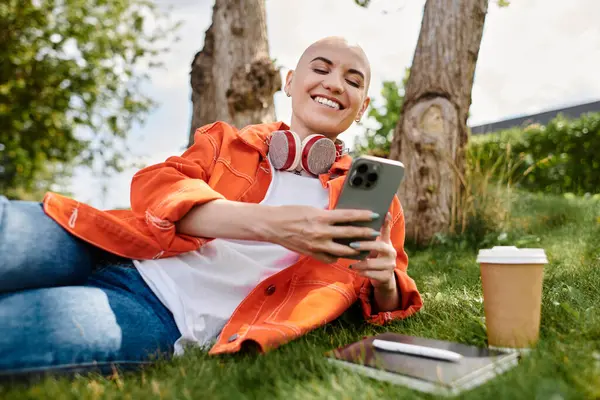 Eine fröhliche junge Frau entspannt sich im Gras und scrollt mit Kopfhörern auf ihrem Handy herum.. — Stockfoto