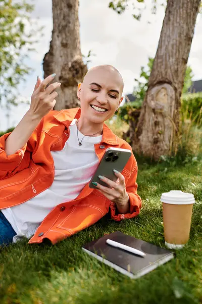 A joyful bald woman sits on lush grass, enjoying her phone and coffee under clear skies. — Stock Photo