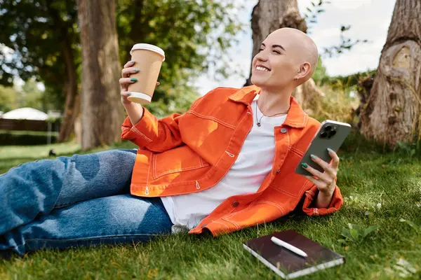 A joyful young woman relaxes on the grass, savoring coffee while using her smartphone. — Stock Photo