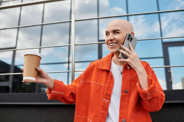 A young bald woman with a warm smile holds a coffee cup and talks animatedly on her phone. — Stock Photo