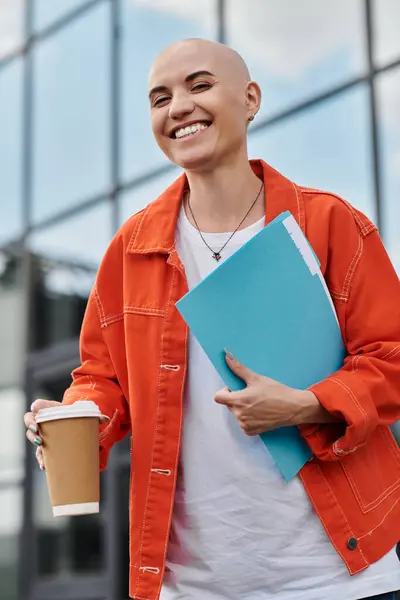 The bald woman radiates joy, clutching a drink and vibrant folders under the clear sky. — Stock Photo