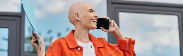A bald woman joyfully chats on her phone, holding a folder in a vibrant orange jacket. — Stock Photo