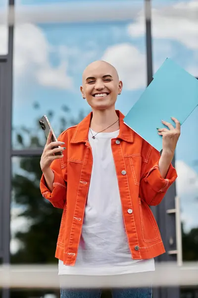 A joyful bald woman holds papers and a phone, radiating confidence by a modern glass structure. — Stock Photo