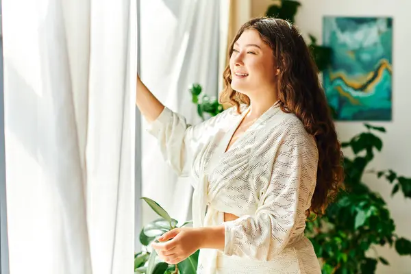 A cheerful young woman with beautiful curly hair brightens her space by opening curtains. — Stock Photo