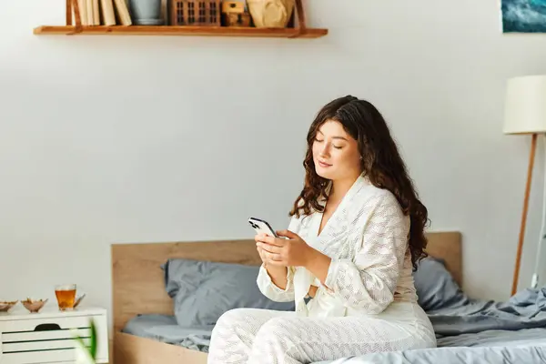A young plus size woman sits comfortably on her bed, engaging with her phone in a cozy atmosphere. — Stock Photo