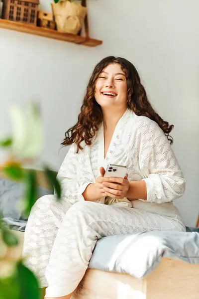 Una joven mujer de talla grande se relaja en su cama, sonriendo brillantemente mientras usa su teléfono inteligente. — Stock Photo