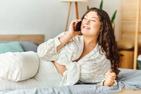 Una mujer alegre de talla grande se relaja en casa, profundamente comprometida en una llamada telefónica en su sofá. - foto de stock