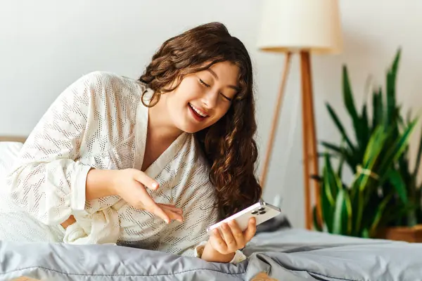 A delighted young woman relaxes on her bed, engaging with her smartphone and smiling. — Stock Photo