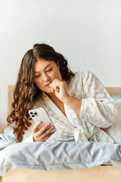 A young beautiful plus size woman lies on her bed, wrapped in a soft robe, engrossed in her phone. — Stock Photo