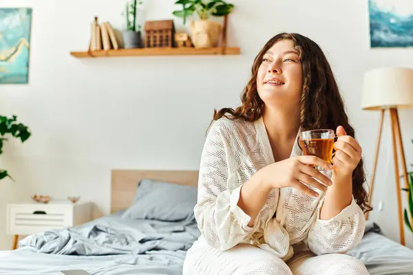 A young plus size woman enjoys a warm cup of tea while relaxing in her cozy bedroom. — Stock Photo