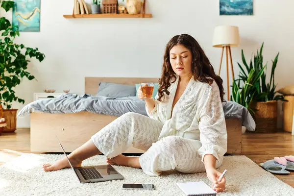 The young woman sits comfortably on the floor, sipping tea and jotting notes as she works. — Stock Photo