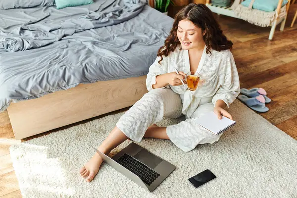 A young woman in a cozy robe sits on the floor, savoring tea and jotting down notes. — Stock Photo