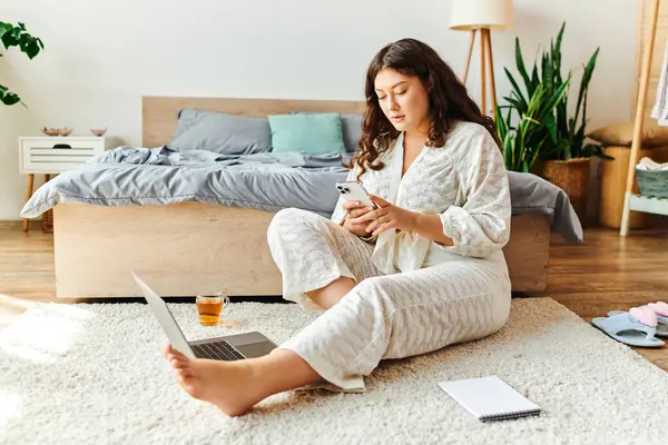 A young beautiful plus size woman unwinds at home, focused on her phone and laptop setup. — Stock Photo