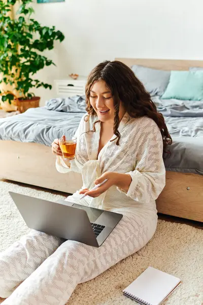 Una giovane bella donna plus size si rilassa a casa, sorseggiando tè mentre si concentra sul suo computer portatile. — Foto stock