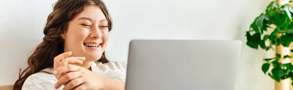 A happy plus size woman smiles as she uses her laptop in a cozy space. — Stock Photo
