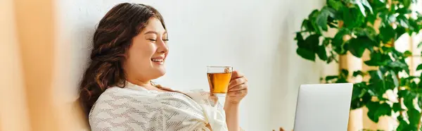 A cheerful plus size woman relaxes at home, sipping tea and engaging with her laptop. — Stock Photo