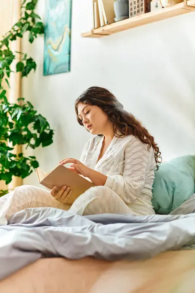 A young beautiful plus size woman relaxes at home, lost in a book while seated on her bed. — Stock Photo