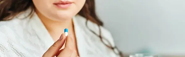 A young plus size woman at home reflects while holding a blue and white capsule in her hand. — Stock Photo