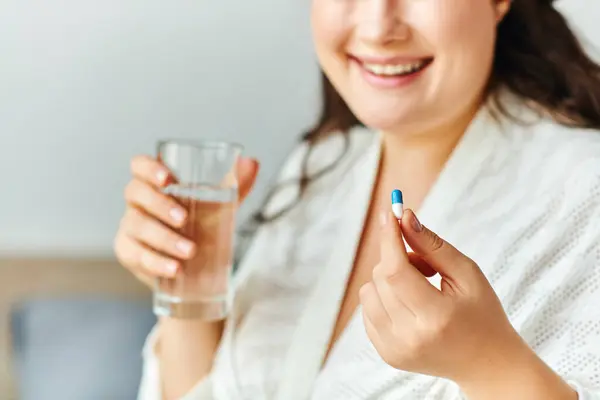 A joyful plus size woman holds a glass of water and a pill, embracing her wellness routine. — Stock Photo
