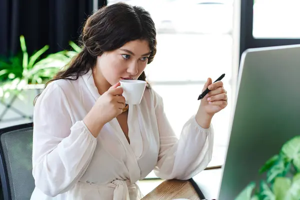 Una giovane bella donna assapora il suo tè mentre si concentra sul suo lavoro al computer in ufficio. — Foto stock