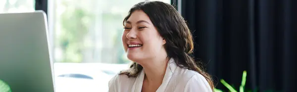A cheerful young woman smiles confidently while engaging with her laptop at office. — Stock Photo