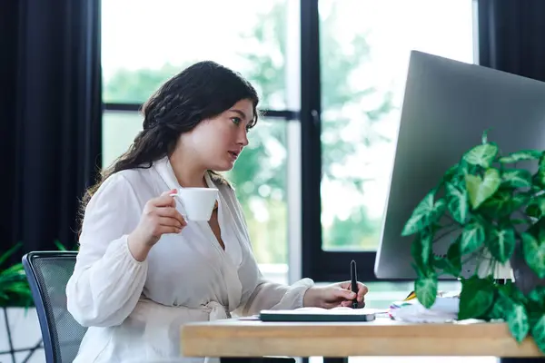 Una giovane bella donna plus size siede alla sua scrivania, sorseggiando caffè mentre lavora sul suo computer portatile. — Foto stock