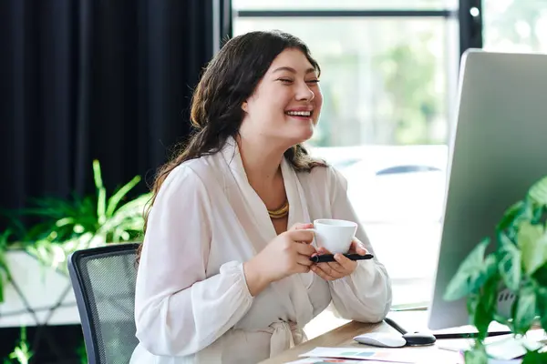A cheerful young woman relaxes at office, sipping coffee while working on her computer. — Stock Photo