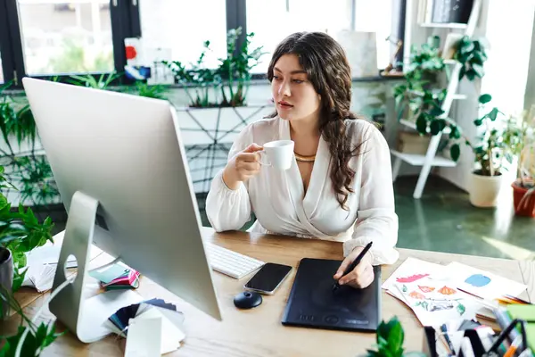 The young woman sips coffee while focusing on her creative work in a bright, plant filled room. — Stock Photo