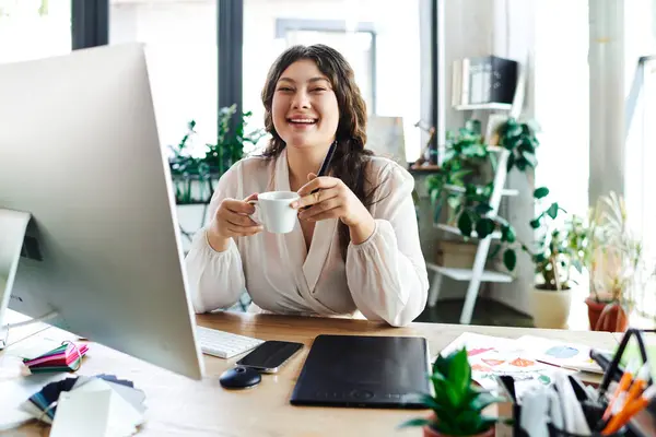 A cheerful young woman sips coffee at her desk, embracing the comfort of her office. — Stock Photo